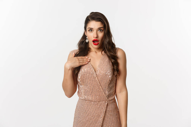 Winter holidays and party concept. Image of attractive woman with red lips, wearing glamour dress and pointing at herself with shock, looking surprised, standing over white background - Foto, Imagem
