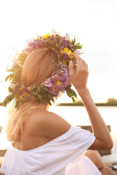 Young woman wearing wreath made of beautiful flowers near river on sunny day - Фото, зображення