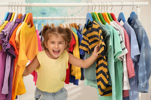 Funny little girl choosing clothes on rack indoors - Foto, imagen
