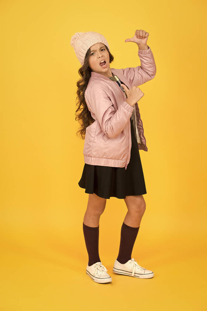 Self confidence concept. Happy child yellow background. Warm clothes for cold season. Fashionable girl looks like hipster. Small girl winter hat. Trendy schoolgirl wear jacket skirt and long socks - Photo, image