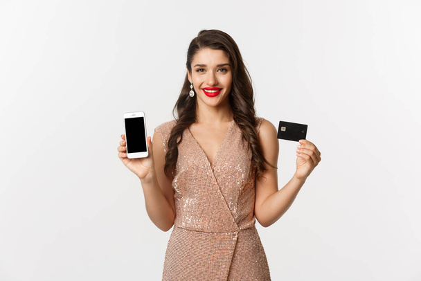 Концепция онлайн-шопинга и праздников. Привлекательная женщина в вечернем платье показывает кредитную карту и мобильный экран, улыбаясь счастливый, белый фон - Фото, изображение