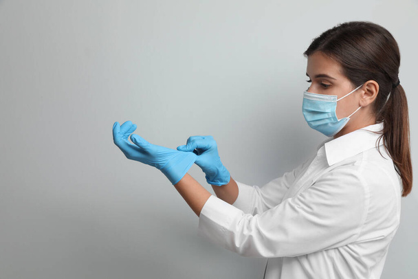 Doctor in protective mask putting on medical gloves against light grey background - Photo, Image