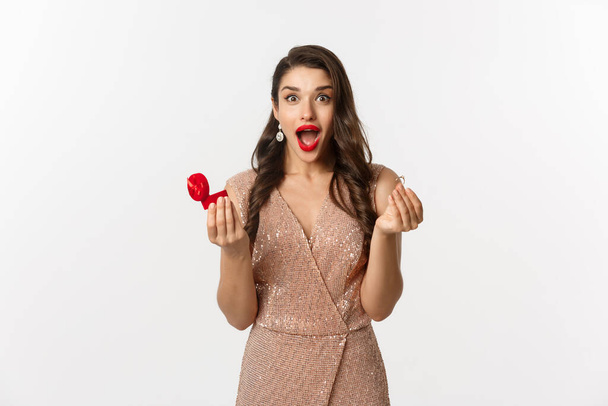 Excited woman holding engagement ring and red box, looking surprised and amazed, receive marriage proposal on date, wearing elegant dress, standing over white background - Photo, image