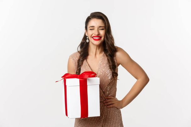 Καλά Χριστούγεννα. Εικόνα της ελκυστικής γυναίκας με πολυτελές φόρεμα, γιορτάζει τις χειμερινές διακοπές, κρατώντας δώρο και γελώντας από την ευτυχία, στέκεται πάνω από το λευκό φόντο - Φωτογραφία, εικόνα