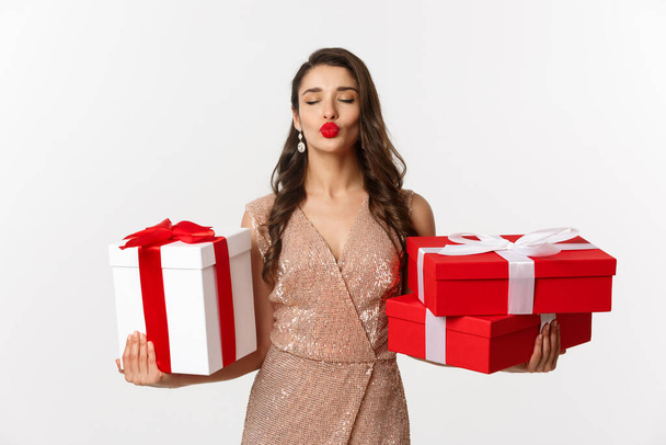 Vacances, concept de fête. Belle femme caucasienne en robe élégante tenant des cadeaux de Noël et souriant heureux, debout sur fond blanc - Photo, image