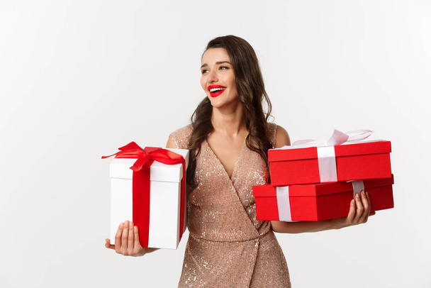 Holidays, celebration concept. Elegant woman with red lips, luxury dress, holding Christmas presents and smiling, enjoying New Year eve, white background - Photo, Image
