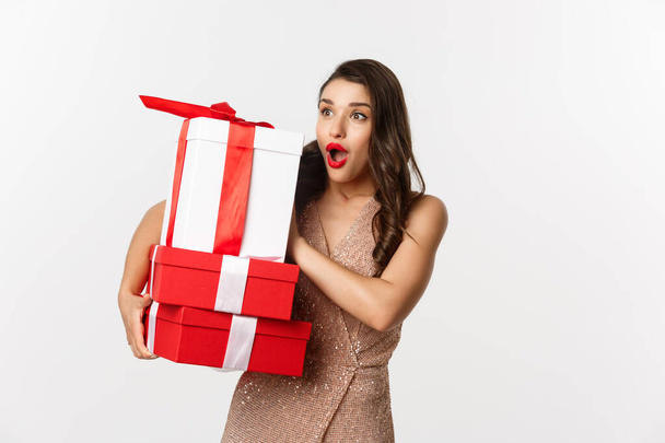 New Year, Christmas and celebration concept. Image of beautiful woman in luxury dress, holding holiday gifts and looking left surprised, standing over white background - Photo, Image