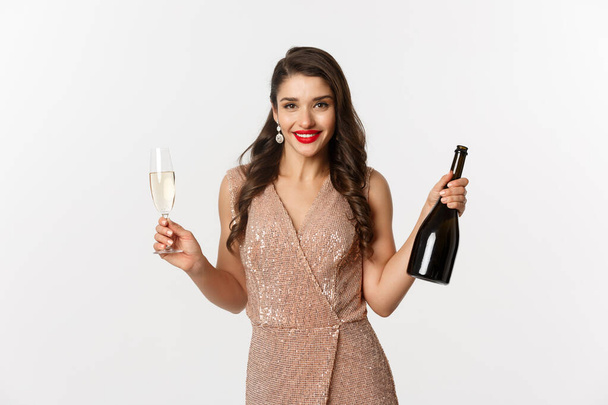 Kış bayramı kutlaması. Lüks elbiseli zarif bir kadın, yeni yıl partisinde içiyor, elinde bardak ve bir şişe şampanya, beyaz arka planda duruyor. - Fotoğraf, Görsel
