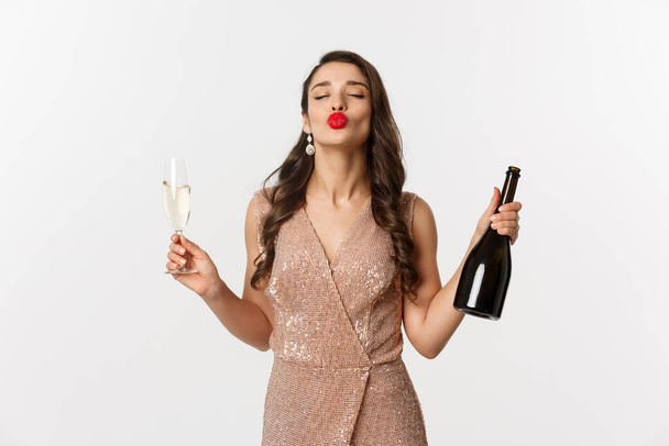 Koncepcja świąt zimowych. Piękna i głupia kobieta w eleganckiej sukience, ciesząca się przyjęciem noworocznym, pijąca szampana, pucker lips for kiss, stojąca nad białym tłem - Zdjęcie, obraz