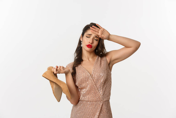 Party and celebration concept. Tired woman in elegant dress take off pair of heels, holding hand on forehead, express fatigue, standing over white background - Foto, Imagem