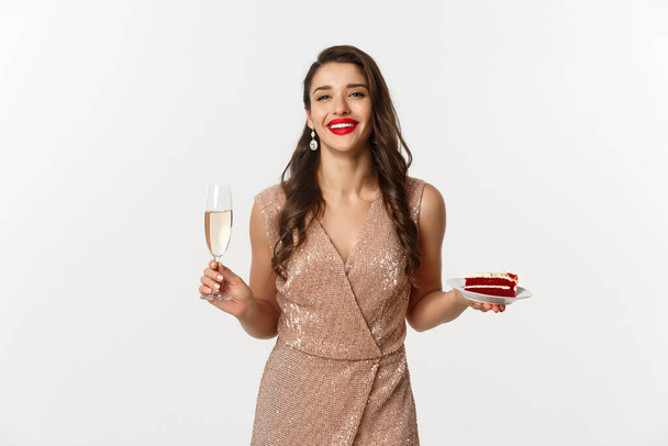 Párty a oslavy. Elegantní žena s červenými rty, půvabné šaty, pití šampaňského a jíst dort, stojící nad bílým pozadím - Fotografie, Obrázek