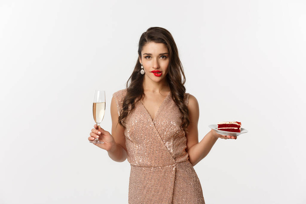 Parti ve kutlama konsepti. Zarif elbiseli seksi bir kadın, elinde şampanya ve bir dilim pasta, dudaklarını ısırıyor ve kameraya çekici görünüyor. - Fotoğraf, Görsel