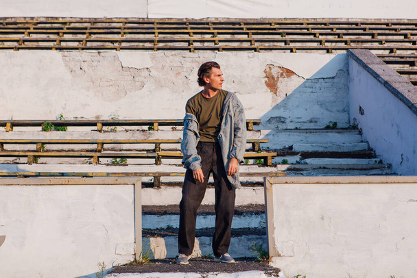 Fashion portrait of a tall handsome man dressed in jeans jacket on the crushed tribune of the old stadium during sunset - Foto, afbeelding