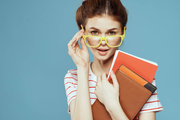 Женщина-студентка с книгами в руках на синем фоне и в желтых очках модель прически обрезанный вид - Фото, изображение