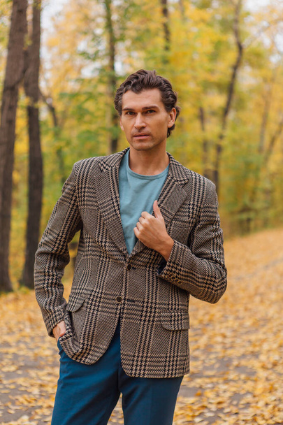 Tall handsome man dressed in a brown jacket walking on the autumn alley - Photo, image