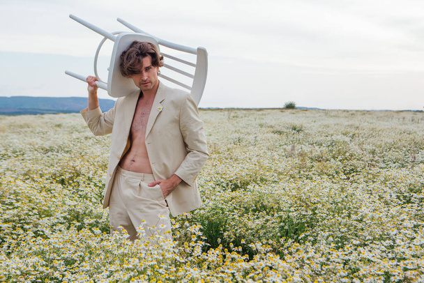 Tall handsome man dressed in a white suit, standing in the middle of camomile flowers field and holding white chair on his shoulder - Zdjęcie, obraz