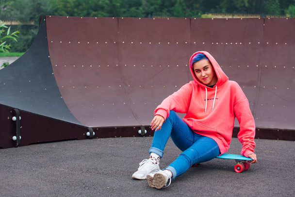 Portrait of a trendy pretty young girl dressed in pink hoodie and jeans sitting next to the skateboard court on her blue plastic skateboard. - Photo, Image