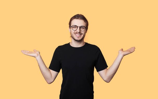 Caucasian man with beard and glasses is comparing two things on his palm while smiling on a yellow studio wall - Foto, afbeelding