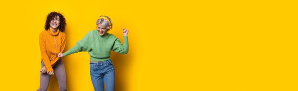 Two cheerful curly haired sisters are listening to music and smile on a yellow studio wall with free space using headphones - Fotó, kép