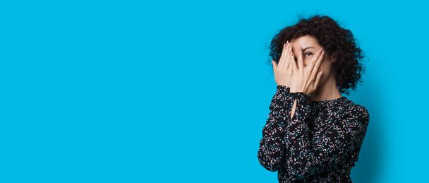 Curly mulher cabelos está cobrindo seu rosto com palmas e sorrir para a câmera posando em uma parede de estúdio azul com espaço livre - Foto, Imagem