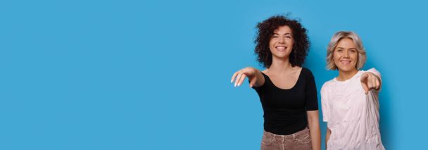 Entzückende Frauen in legerer Kleidung zeigen lächelnd in die Kamera an einer blauen Studiowand mit freiem Raum - Foto, Bild