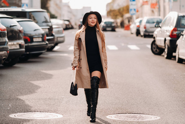 Stijlvolle jonge vrouw in een beige jas met een zwarte hoed op een straat. Straatmode voor vrouwen. Herfst kleding. Stedelijke stijl. - Foto, afbeelding