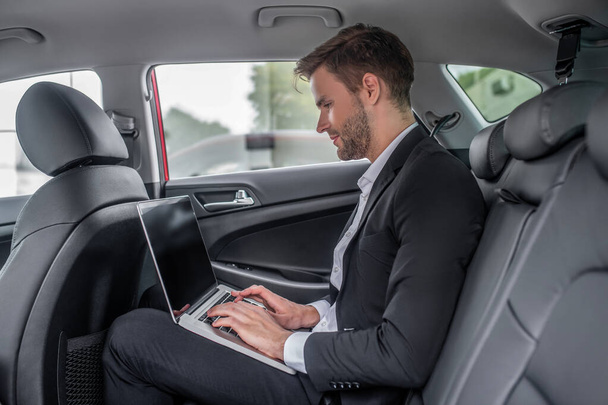 Brown-haired male sitting at backseat of car, typing on laptop - Фото, зображення