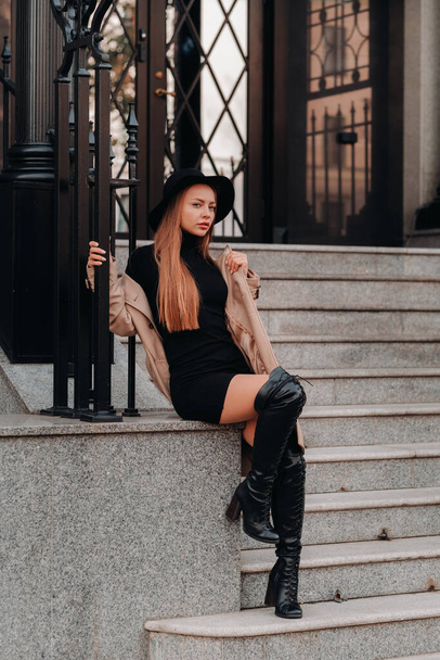 Stylish young woman in a beige coat in a black hat on a city street. Women's street fashion. Autumn clothing.Urban style. - Φωτογραφία, εικόνα