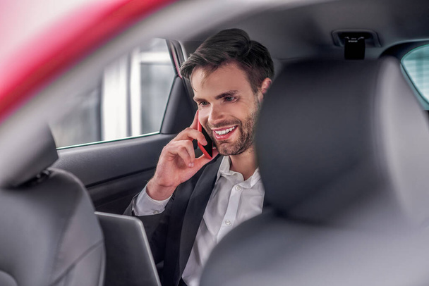Smiling male sitting at backseat of car with laptop, talking on phone - Фото, изображение