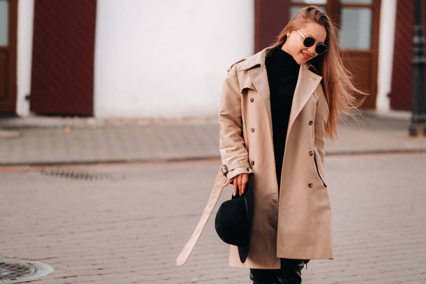 Stylish young woman in a beige coat and a black hat in his hands and glasses on a city street. Women's street fashion. Autumn clothing.Urban style - Foto, Imagem
