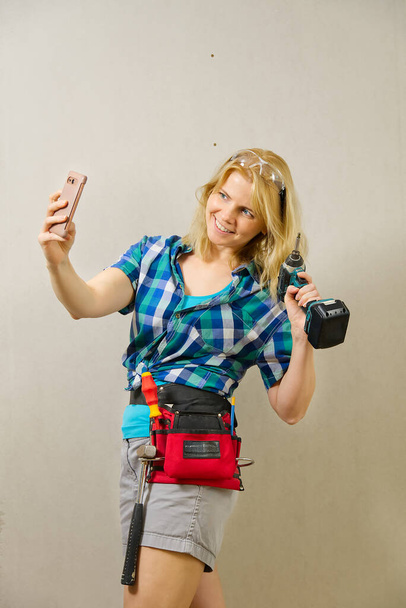 Blond vrouwtje met een doe-het-zelvers gereedschapsriem vol met verschillende gereedschappen op een onbeschilderde gipsplaten muurachtergrond. DIY vrouw concept. - Foto, afbeelding