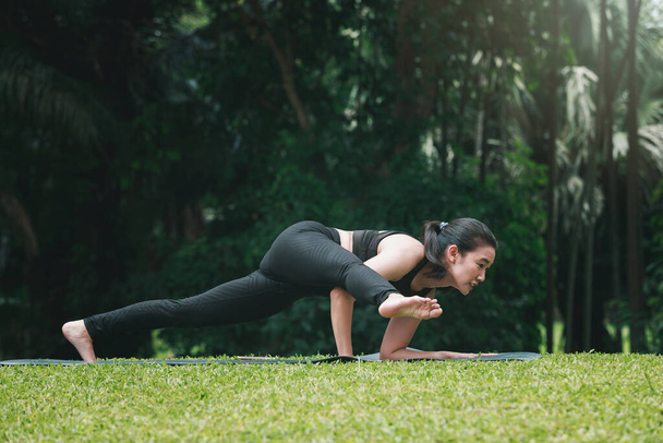 Donna asiatica che pratica yoga in posa Dedicato al saggio Koundinya II (Eka Pada Koundinyanasana II) sul tappeto nel parco all'aperto. - Foto, immagini