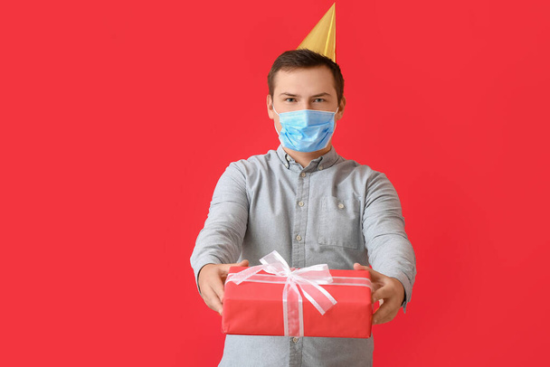 Young man in medical mask and with Birthday gift on color background - Foto, imagen