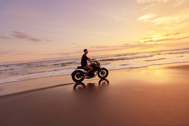 Mann und Motorrad am Ocean Beach bei schönem tropischen Sonnenuntergang. Schöner Biker auf dem Motorrad an der Sandküste in Bali, Indonesien. - Foto, Bild