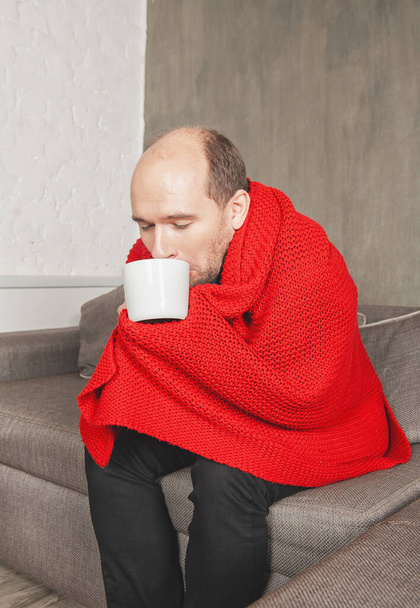Sick man with cup of hot drink wrapped in warm knitted red plaid sneezes at hom - Foto, Imagen