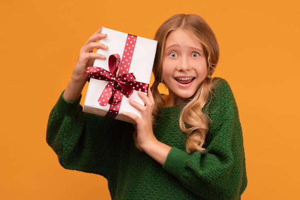 Imagen de la encantadora chica rubia de 12-14 años en suéter verde cálido sonriendo y sosteniendo la caja actual con lazo rojo. Fotografía del estudio, fondo amarillo. Año Nuevo Día de la Mujer Cumpleaños Concepto de vacaciones - Foto, Imagen