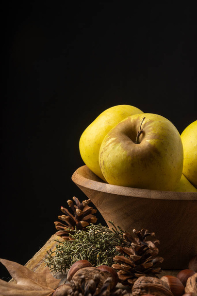 Primer plano de manzanas amarillas en cuenco de madera sobre mesa de madera con frutos secos y motivos otoñales, fondo negro vertical - Foto, Imagen