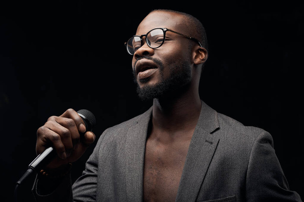 portrait of a dark-skinned handsome guy in grey jacket and glasses  on a black background, singing in a microphone emotionally - Photo, image