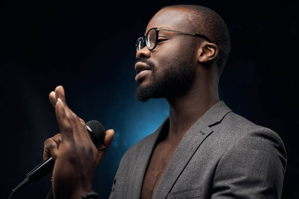 portrait of a dark-skinned handsome guy in grey jacket and glasses  on a dark blue background, singing in a microphone  with closed eyes  - Фото, изображение