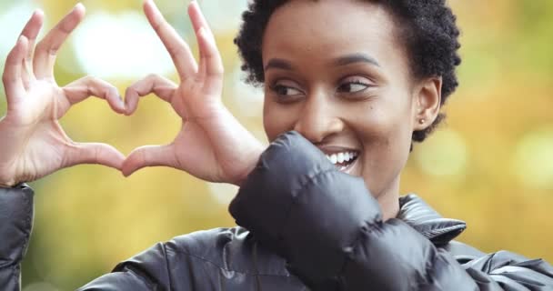 Porträt lächelnd emotional freundlich ethnisch-afrikanisch-amerikanische Frau, die im Herbst draußen steht und Hände zeigt, Symbol der Liebe Zeichen der Friedensromantik macht Finger Herzform lächelnden Blick in die Kamera - Filmmaterial, Video