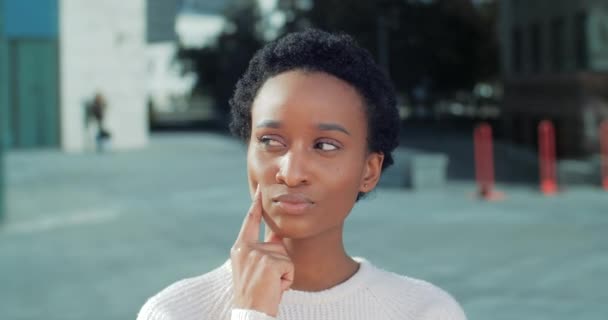 Portret van een Afrikaanse vrouw in het wit die in de buitenlucht staat in de buurt van het stadsgebouw zet haar vinger onder ogen te herinneren denken over het idee. Hoofd schot van broeden pensive gemengde ras etnische meisje student planning - Video
