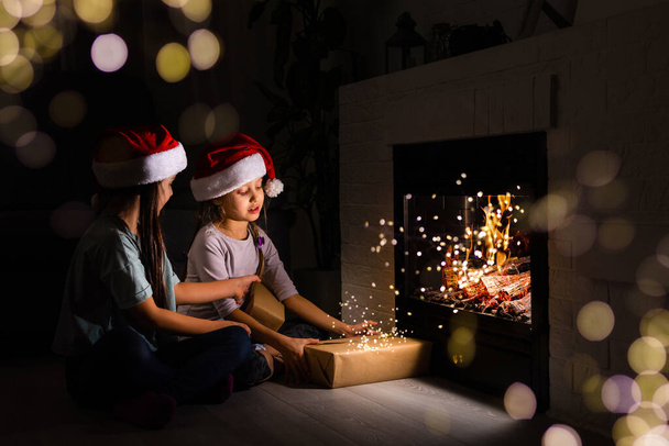 two little girls sitting by a fireplace in a cozy dark living room on Christmas eve - Photo, Image
