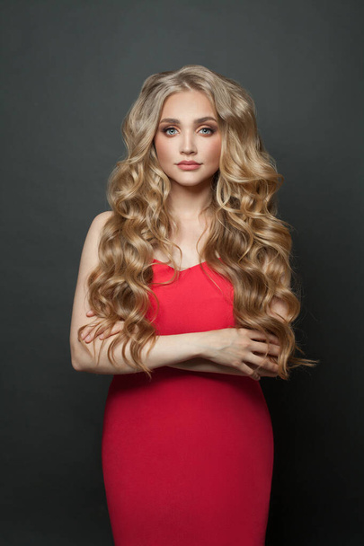 Beautiful fashion model woman with long blonde curly hair wearing red evening gown on black background - Foto, imagen