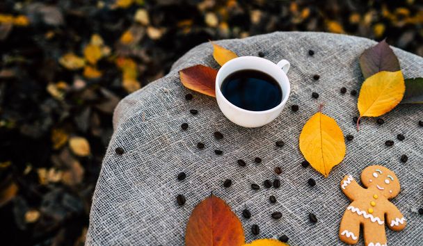 Gingerbread man and cup of coffee near autumn leaves. Top view of sweet gingerbread man and cup of strong espresso coffee placed near colorful autumn leaves on linen cloth in park. - Foto, afbeelding