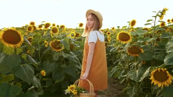 Una mujer con una cesta de flores está mirando a la cámara y sonriendo. Está de pie entre los girasoles. 4K - Metraje, vídeo