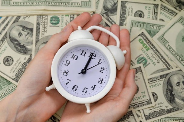 Женские руки с белым будильником лежат на фоне долларовых купюр крупным планом. Концепция финансов, бизнеса - Фото, изображение