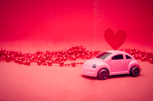Small pink car with a red heart on the roof of the car on a red background with beads with copy space. Festive bright background - Photo, image