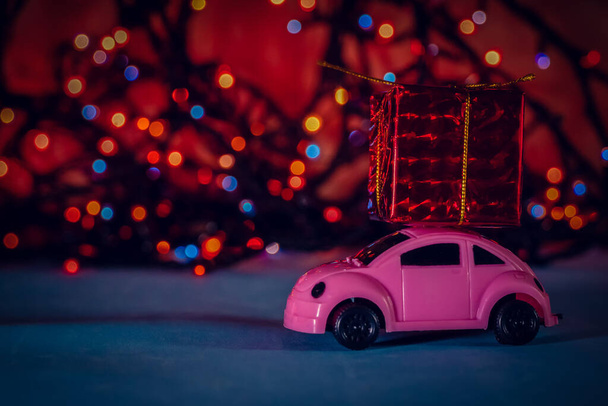 Могилев, Беларусь - 10 сентября 2020 года: Розовый игрушечный автомобиль с красным подарком на крыше на фоне рождественской гирлянды на синем фоне с копировальным местом. Праздничный красивый новогодний фон - Фото, изображение