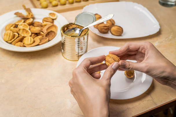 Τα χέρια των γυναικών συνδέουν τα μισά του μπισκότου Nut γεμισμένο με βραστό συμπυκνωμένο γάλα πάνω από το τραπέζι με πιάτα, μπάλες ζύμης μπισκότων, έτοιμα μπισκότα μισά, ένα βάζο βραστό συμπυκνωμένο γάλα - Φωτογραφία, εικόνα