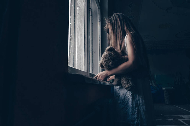 dívka v bílých šatech s dlouhými vlasy a medvídkem pod paží stojí u okna a čte knihu ve tmě. Pojem horor, mysticismus - Fotografie, Obrázek
